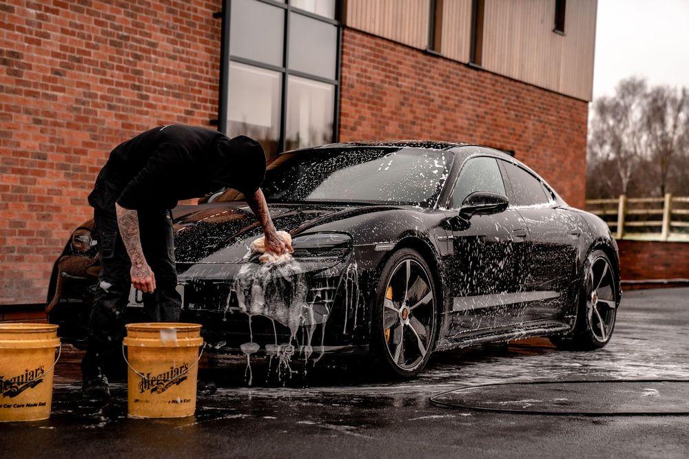 Person hand washing black car by brick building.