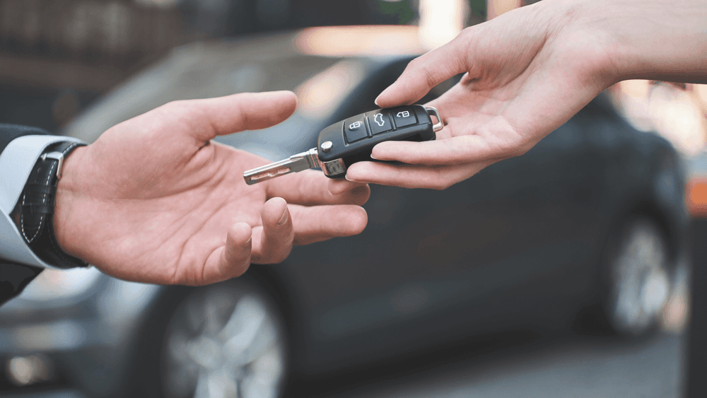Woman's hand holding a key and handing it over to a man's hand with a car behind them