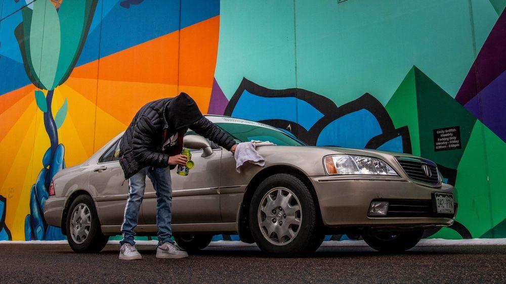 Man in a hoodie and jacket polishing his car in front of colorful wall.
