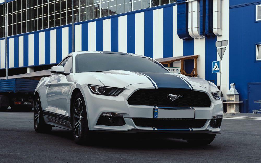White Ford Mustang with blue stripe in front of white and blue building.