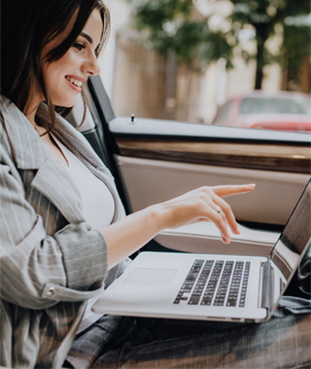 A smiling woman sitting in the driver's seat of a car, pointing at the screen of a laptop resting on her lap